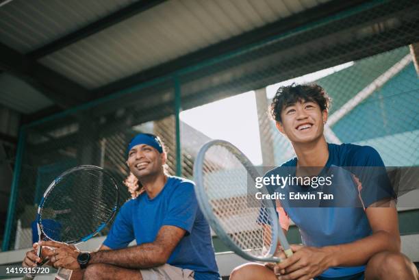 tenista profesional asiático sonriendo sentado en el estadio observando a sus compañeros de equipo practicando en la cancha de tenis - atuendo de tenis fotografías e imágenes de stock