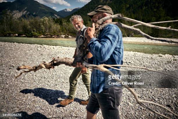 two friends carrying wood at a riverbank - überlebenskünstler stock-fotos und bilder