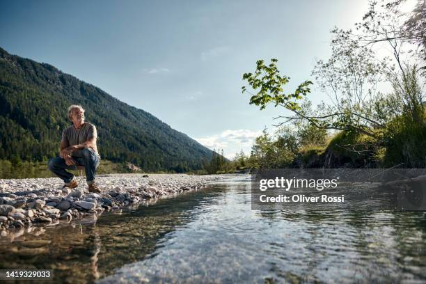 mature man squatting  at a riverbank - riverbank stockfoto's en -beelden