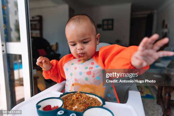 angry 7 month old baby eating food from a plate. dressed with a bib and sitting on a highchair. - よだれ掛け ストックフォトと画像
