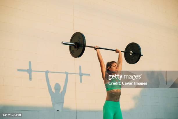 young active woman doing overhead press with barbell in morning - lange halter stockfoto's en -beelden