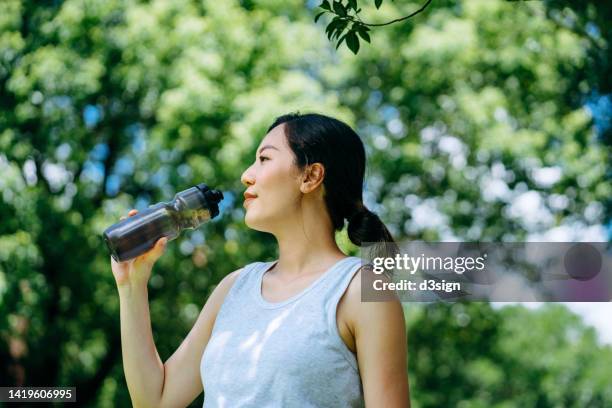 young active asian woman drinking water from reusable water bottle after exercising outdoors in nature park. taking a water break. staying hydrated. health and fitness training routine. healthy living lifestyle - running refreshment stock pictures, royalty-free photos & images