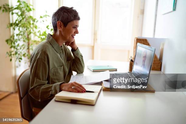 woman following online courses on her laptop at home - mature student stockfoto's en -beelden