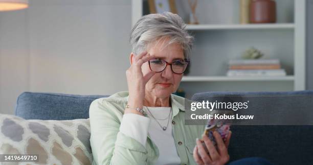 mature woman reading news on phone while relaxing on a sofa at home, carefree and content. senior female using an easy online app, browsing for deal or discount on the internet or renew subscription - subscription stock pictures, royalty-free photos & images