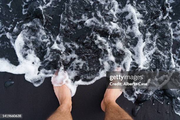 bare feet submerged in sea water on a black volcanic sand beach - mens bare feet fotografías e imágenes de stock