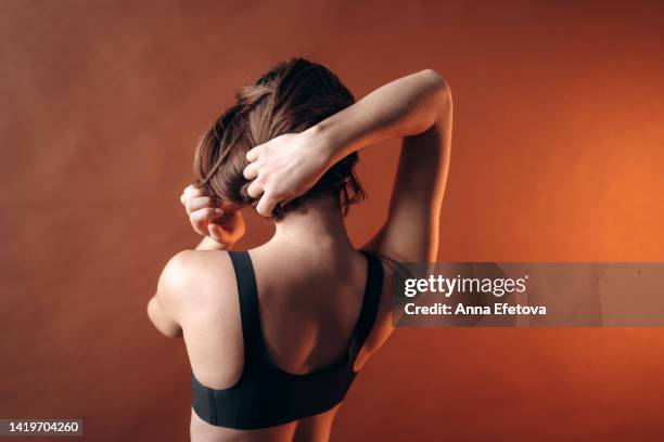 beautiful young woman in black bra and black pants is adjusting her short hair posing against brown wall background. concept of natural beauty. rear view - adjusting hair stock pictures, royalty-free photos & images