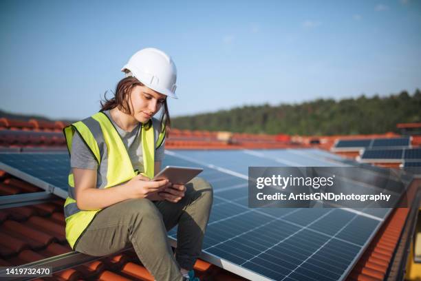 female engineer sitting on a rooftop with solar panels using technology. - solar panel installation stock pictures, royalty-free photos & images
