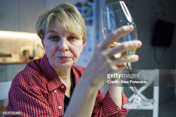 tired mature  blond woman wearing a red shirt is drinking white wine from a wineglass. - unhappy woman blonde glasses stock pictures, royalty-free photos & images