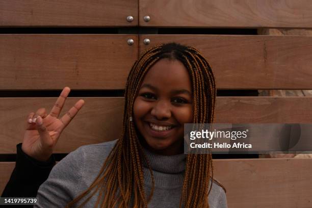 a beautiful young woman smiles radiantly on a background of timber whilst making a peace sign, a teenage girl with a big toothy smile & long braided & beaded brown hair and an expression of total enjoyment makes a v hand sign or gesture for victory - victory sign smile foto e immagini stock