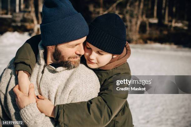 affectionate father and son wearing knit hat embracing each other on sunny day - warm clothing stock pictures, royalty-free photos & images