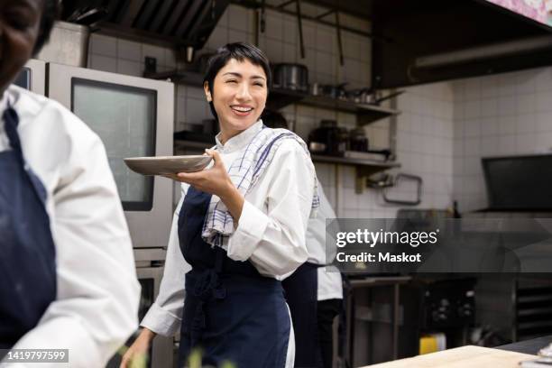 smiling chef carrying plate while walking in restaurant kitchen - gastronom stock-fotos und bilder