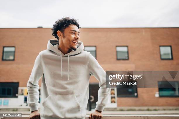 happy young man looking away while standing in front of building - hooded top stock pictures, royalty-free photos & images
