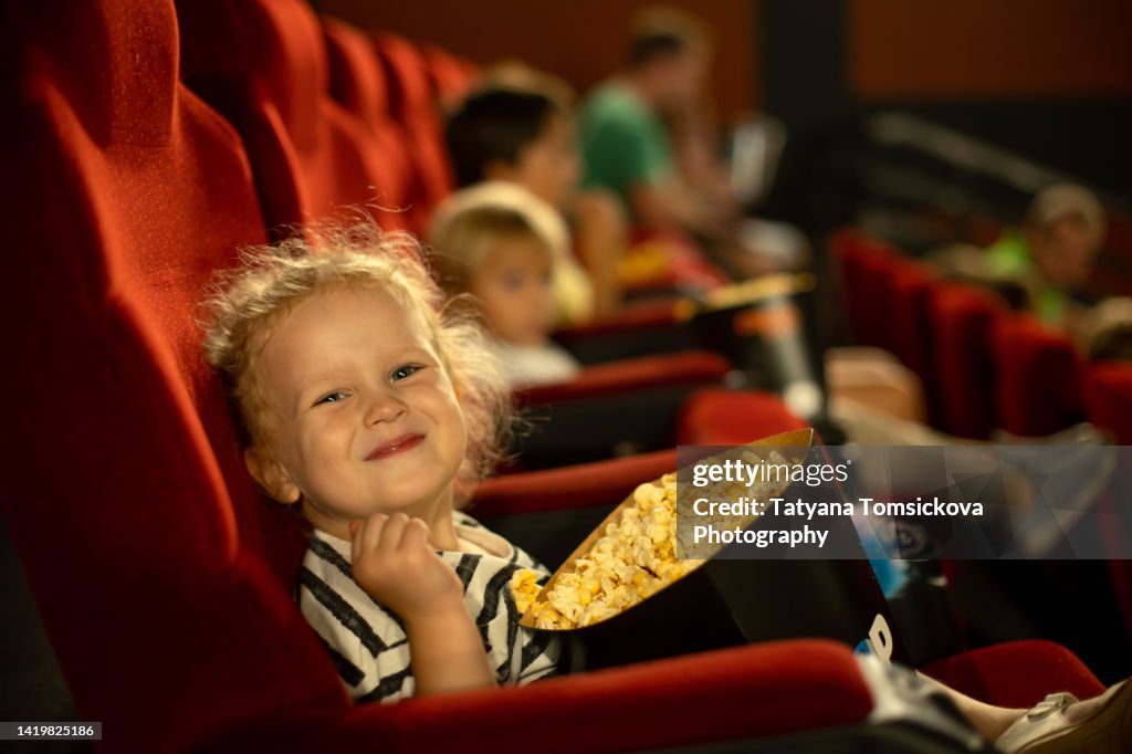 Cute child, curly girl, watching movie in a cinema, eating popcorn and enjoying