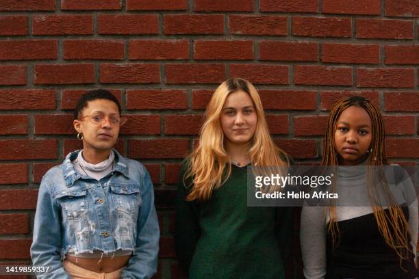 three casually dressed beautiful young women, teenage girls stand in front of a red brick wall & where the friend in the centre has long blond hair & on her girl friends on either side have a short afro & long brown braids - girl band - fotografias e filmes do acervo