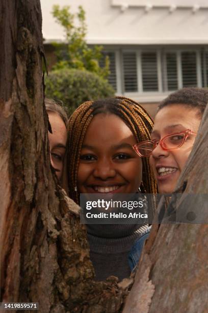 toothy smiles behind a tree, three beautiful young women, teenage girls with different hairstyles in dark afro, straight dyed blond & brown braided peek from behind a tree & smile radiantly at camera - victory sign smile foto e immagini stock