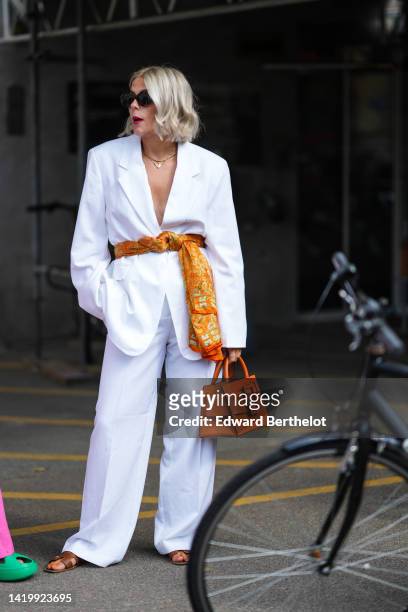 Guest wears black sunglasses, a white oversized blazer jacket, matching white large pants, a yellow and orange print pattern silk scarf as a belt, an...