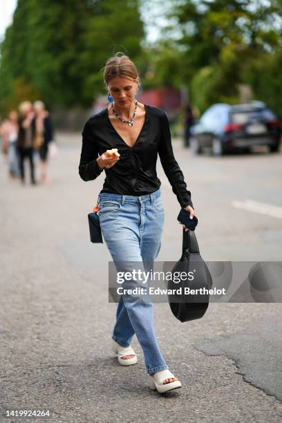 Guest wears blue and gold earrings, a white and blue pearls pendant necklace, a black V-neck / long sleeves / ruffled satin pullover, blue denim wide...