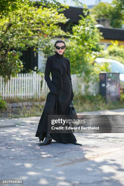 Guest wears black butterfly sunglasses, gold earrings, a black high neck / long sleeves / long dress, a black shiny leather handbag, black shiny...