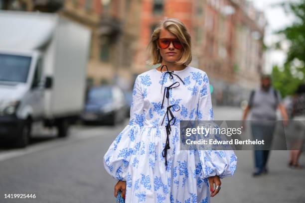 Guest wears red sunglasses, silver earrings, a white with blue print pattern long puffy sleeves / midi dress with black laces neck, silver rings ,...