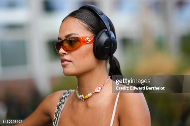 Guest wears black headphones, orange sunglasses from Chanel, a multicolored pearls necklace, a black heart-neck with white borders long dress, a...