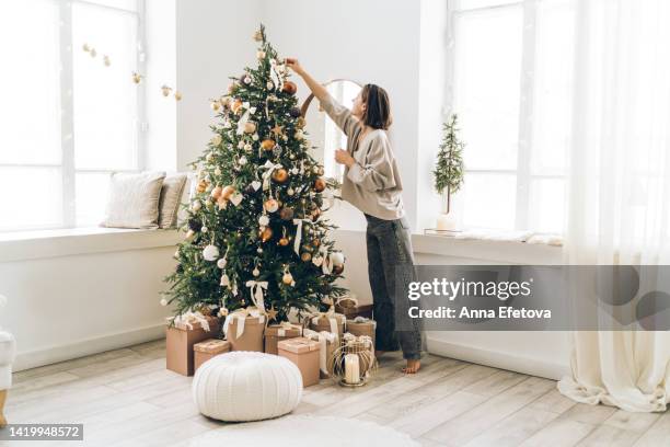 young woman is decorating christmas tree with many different decorations and festive garland in minimalist light interior. new year celebration concept - kerstboom versieren stockfoto's en -beelden