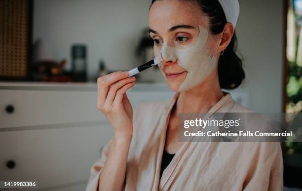 a woman uses a brush to apply a clay face mask to her skin - blackhead stock-fotos und bilder