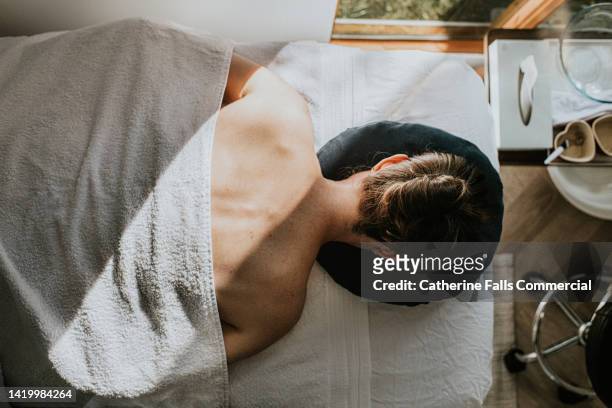 a woman lays face down on a massage table, covered by a towel. her bare shoulders and upper back are visible. - masaje fotografías e imágenes de stock