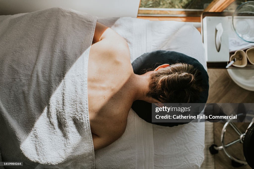 A woman lays face down on a massage table, covered by a towel. Her bare shoulders and upper back are visible.