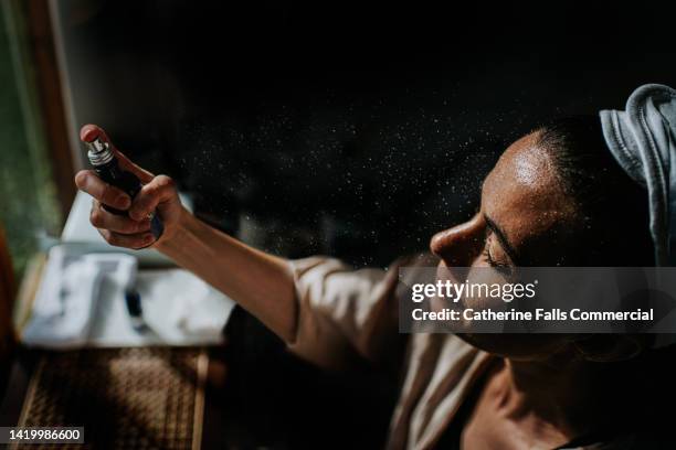 cinematic image of a woman holding a spray bottle, and spritzing moisture on to her face. droplets sit on her face, - cooling down stockfoto's en -beelden