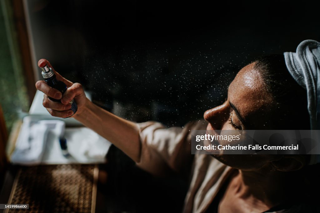 Cinematic image of a woman holding a spray bottle, and spritzing moisture on to her face. Droplets sit on her face,