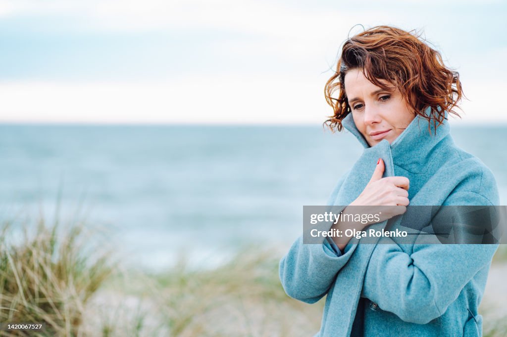 Abandoned, lonely day on beach. Sad mature woman in warm overcoat covering by collar on windy day on beach