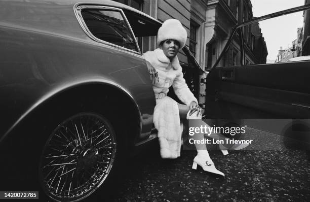 British singer Shirley Bassey wearing a white mink coat and matching hat, with knee-length boots and a ring on every finger, sitting in the driving...