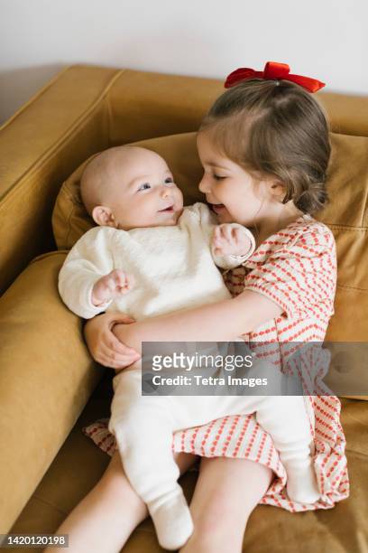 smiling girl (2-3) holding baby brother (2-5 months) brother on sofa - 2 5 months stock pictures, royalty-free photos & images