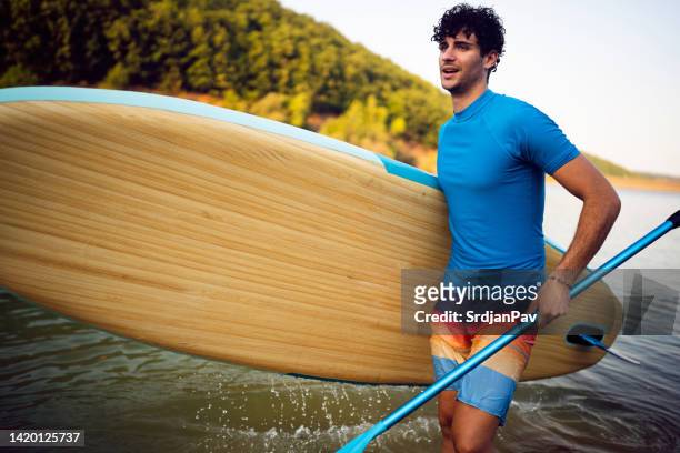 caucasian man, leaving the lake, while carrying standup paddleboard - ventil stockfoto's en -beelden