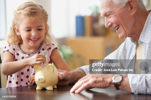 caucasian grandfather watching granddaughter putting coin into piggy bank - spaarpot financieel item stockfoto's en -beelden