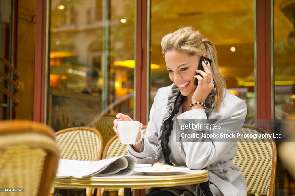 Hispanic woman drinking coffee and using cell phone in outdoor cafe