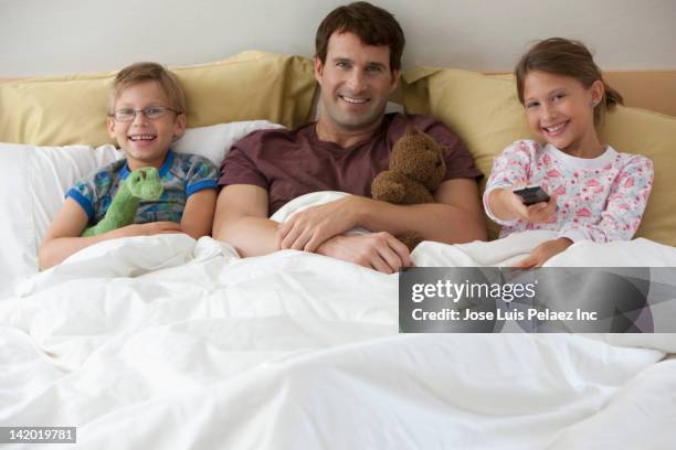 caucasian father and children watching television in bed - three people in bed stock pictures, royalty-free photos & images