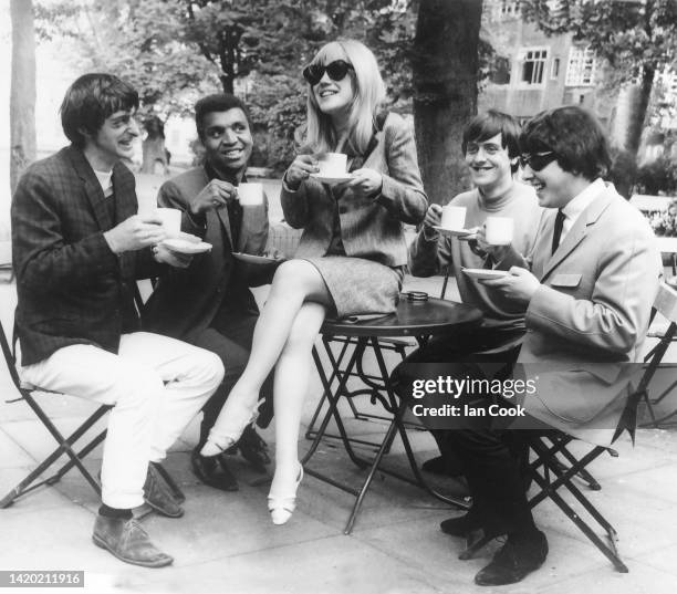 English Pop singer Marianne Faithfull sits on an outdoor table as she drinks a cup of tea with fellow musician Kenny Lynch and unidentified others...