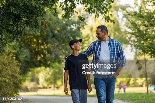 Front view of father and son walking embraced at the park, chatting and bonding Front view of father and son walking embraced at the park, chatting and bonding