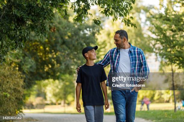 front view of father and son walking embraced at the park, chatting and bonding - teenage son stock pictures, royalty-free photos & images