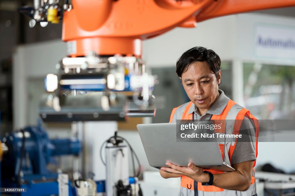 High Precise and Zero-cost Solution for Fully Automatic Industrial Robot. Japanese Engineer uses a laptop computer to configure of hydraulic, electric, or pneumatic drive systems to control gripper fingers in car battery manufacturing processes.