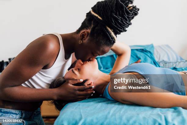 young woman kissing girlfriend lying on bed at home - lesbian love fotografías e imágenes de stock