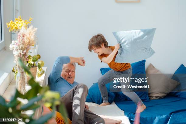 happy boy playing pillow fight with grandfather on sofa at home - pillow fight fotografías e imágenes de stock