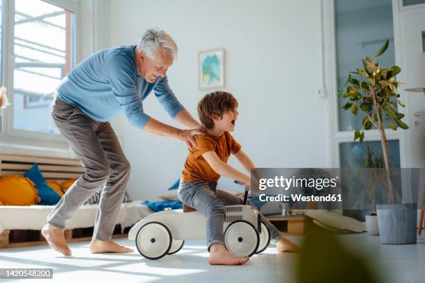 playful senior man pushing grandson sitting on toy car in living room at home - aktiver senior stock-fotos und bilder