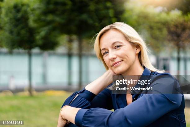 smiling mature businesswoman with hand in blond hair at park - une seule femme d'âge mûr photos et images de collection