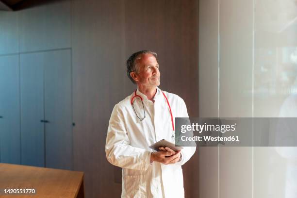 smiling doctor holding tablet pc standing near glass wall in hospital - westend 61 fotografías e imágenes de stock