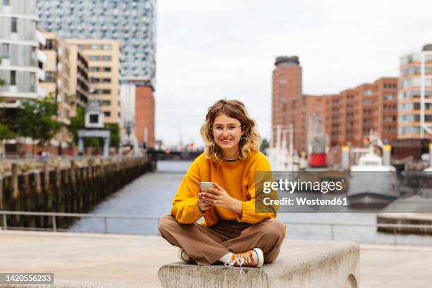 happy woman with smart phone sitting on bench at elbphilharmonie - hamburgo fotografías e imágenes de stock