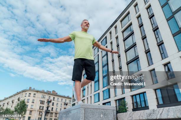 mature man with arms outstretched exercising on concrete by building - auf einem bein stock-fotos und bilder