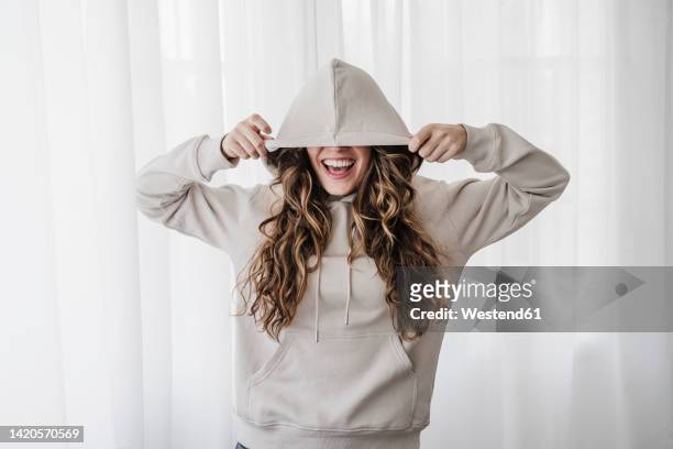 playful woman covering eyes with hood at home - welliges haar stock-fotos und bilder