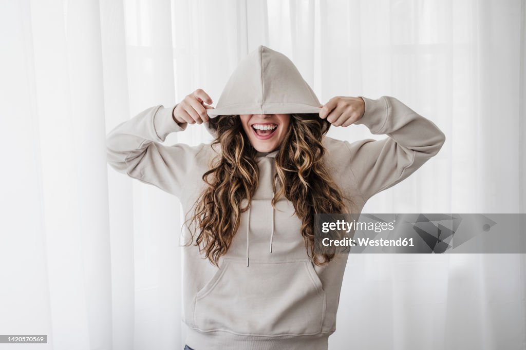 Playful woman covering eyes with hood at home